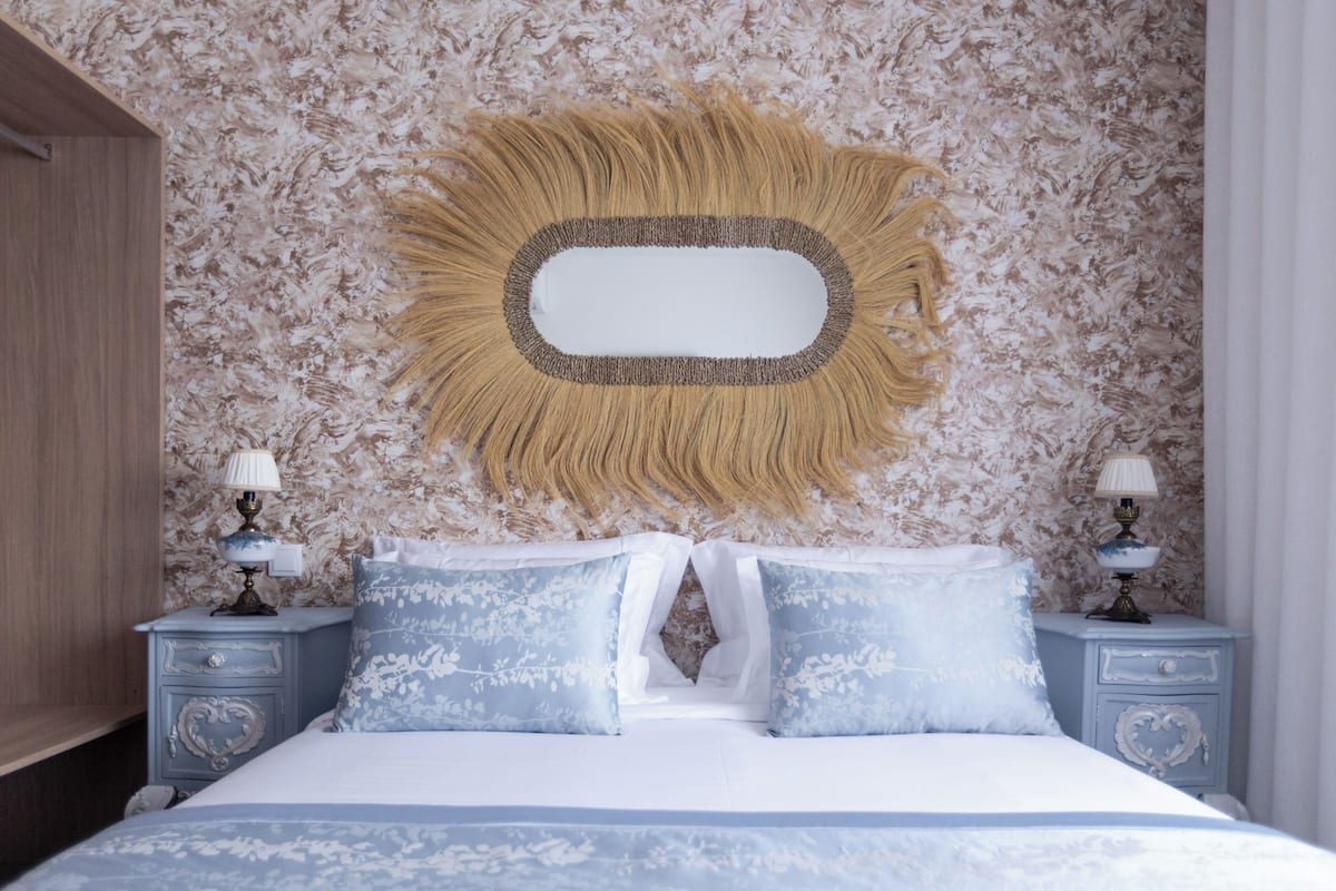 A bedroom with a bed, two nightstands with lamps, a decorative mirror, and patterned wallpaper.