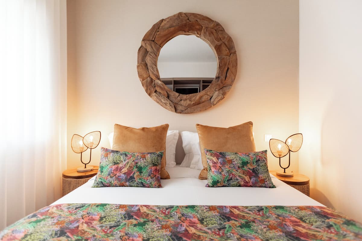A neatly made bed with a colorful floral bedspread, two brown pillows, and a round mirror on the wall above.