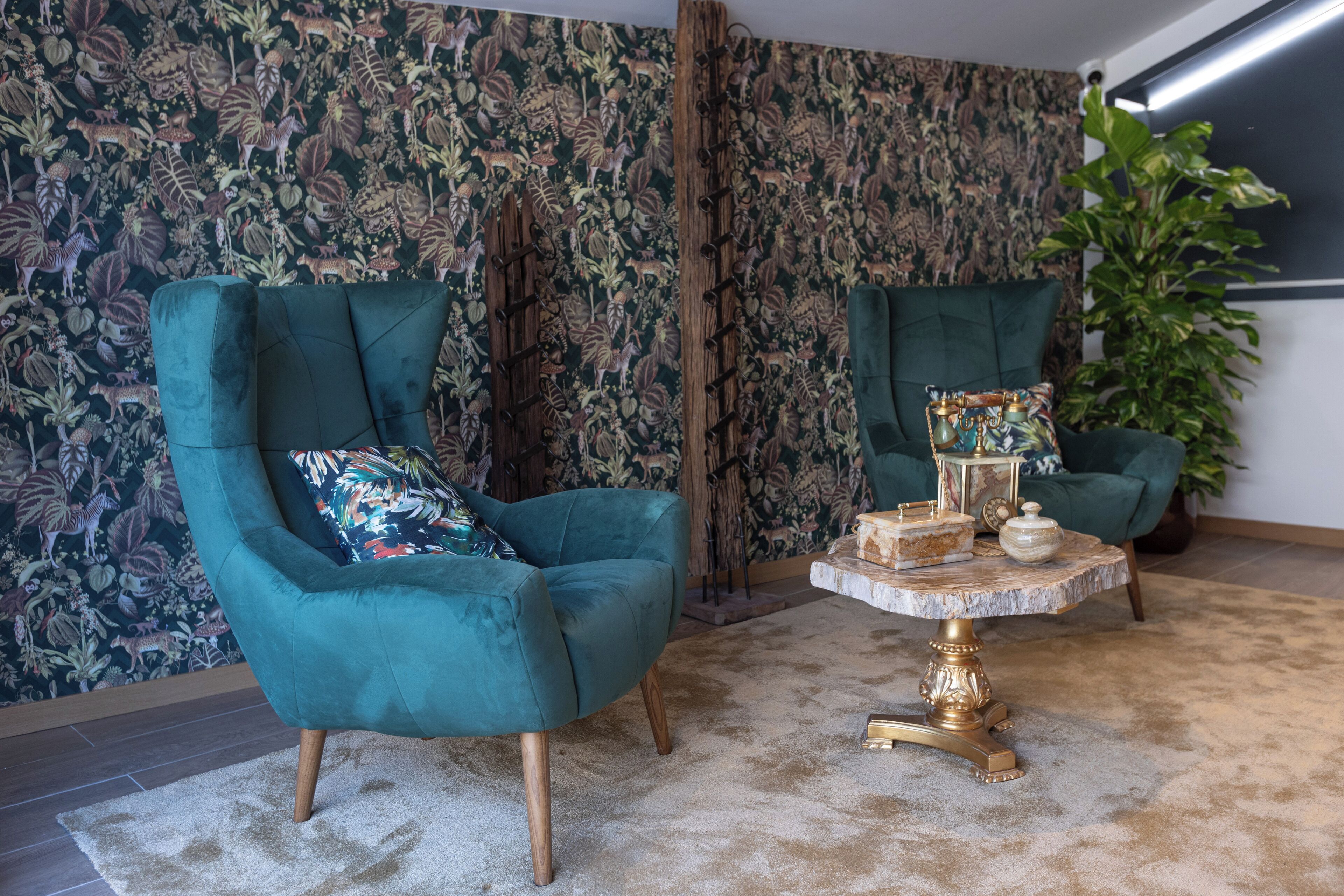 A room with two teal armchairs, a round coffee table with a gold base, and a patterned wallpaper.
