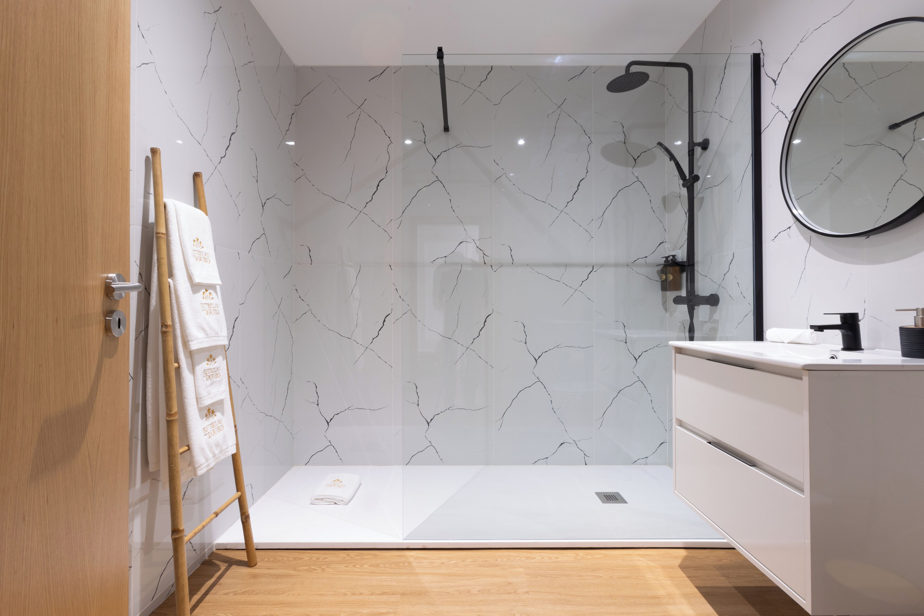 A modern bathroom with a glass shower enclosure, a wooden floor, a white vanity with a mirror, and a wooden door.