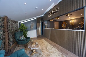 Un vestíbulo moderno con un sillón verde, una mesa de centro redonda y una recepción con paneles de madera oscura y detalles dorados.