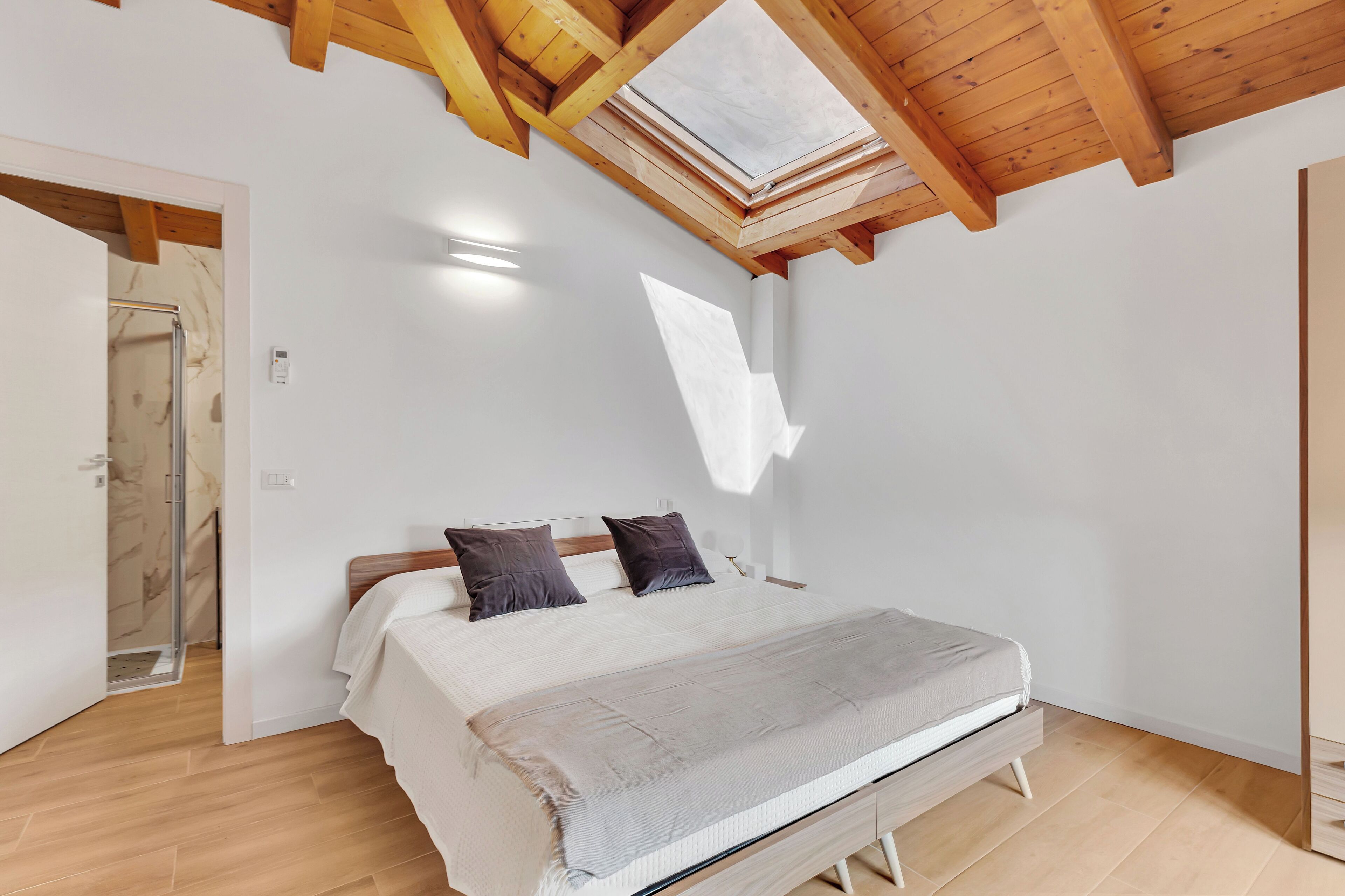 A bedroom with a bed, wooden beams, a skylight, and a door leading to a bathroom.