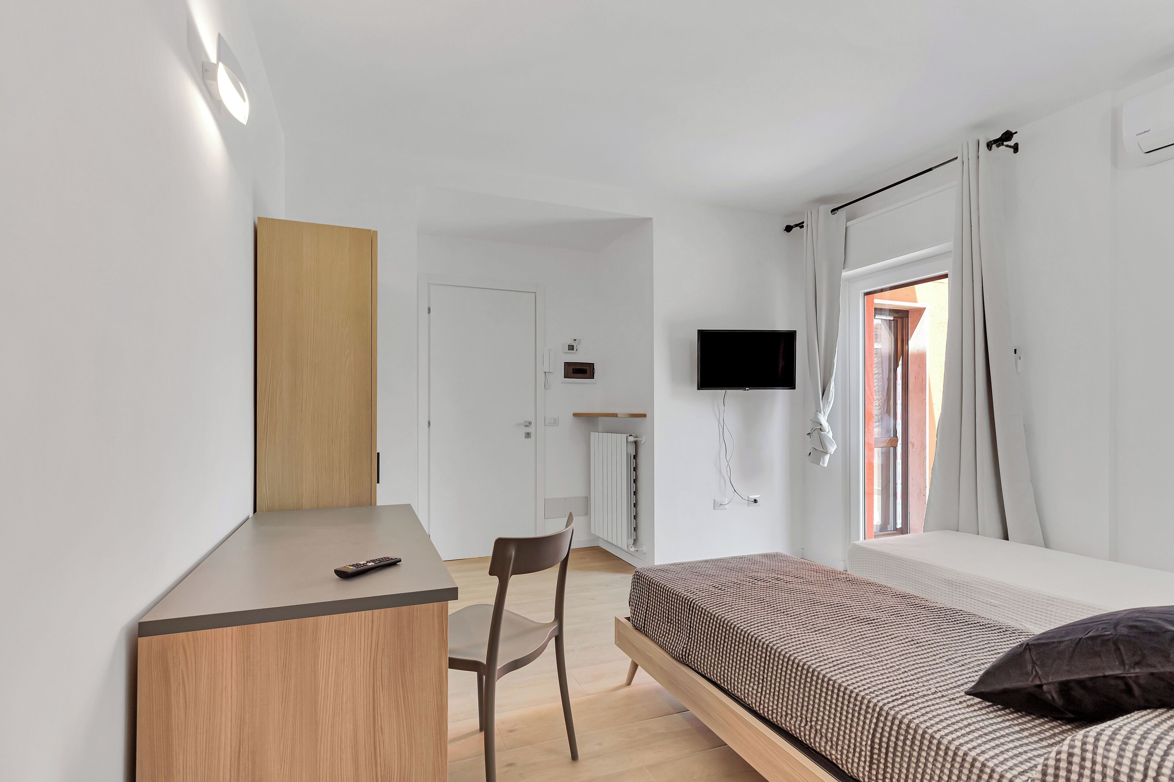 Un dormitorio con cama, escritorio, silla, televisor y ventana con cortinas.