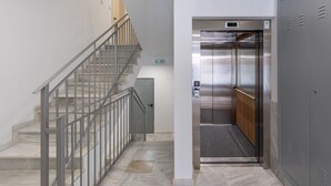 Un ascensor moderno con pantalla digital, una escalera de mármol con barandillas metálicas y un casillero gris a la derecha.
