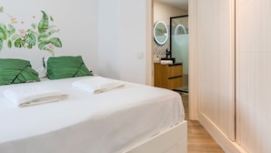 Un dormitorio con una cama, almohadillas verdes a rayas, una colcha blanca, un espejo fijado a la pared y un armario de madera.