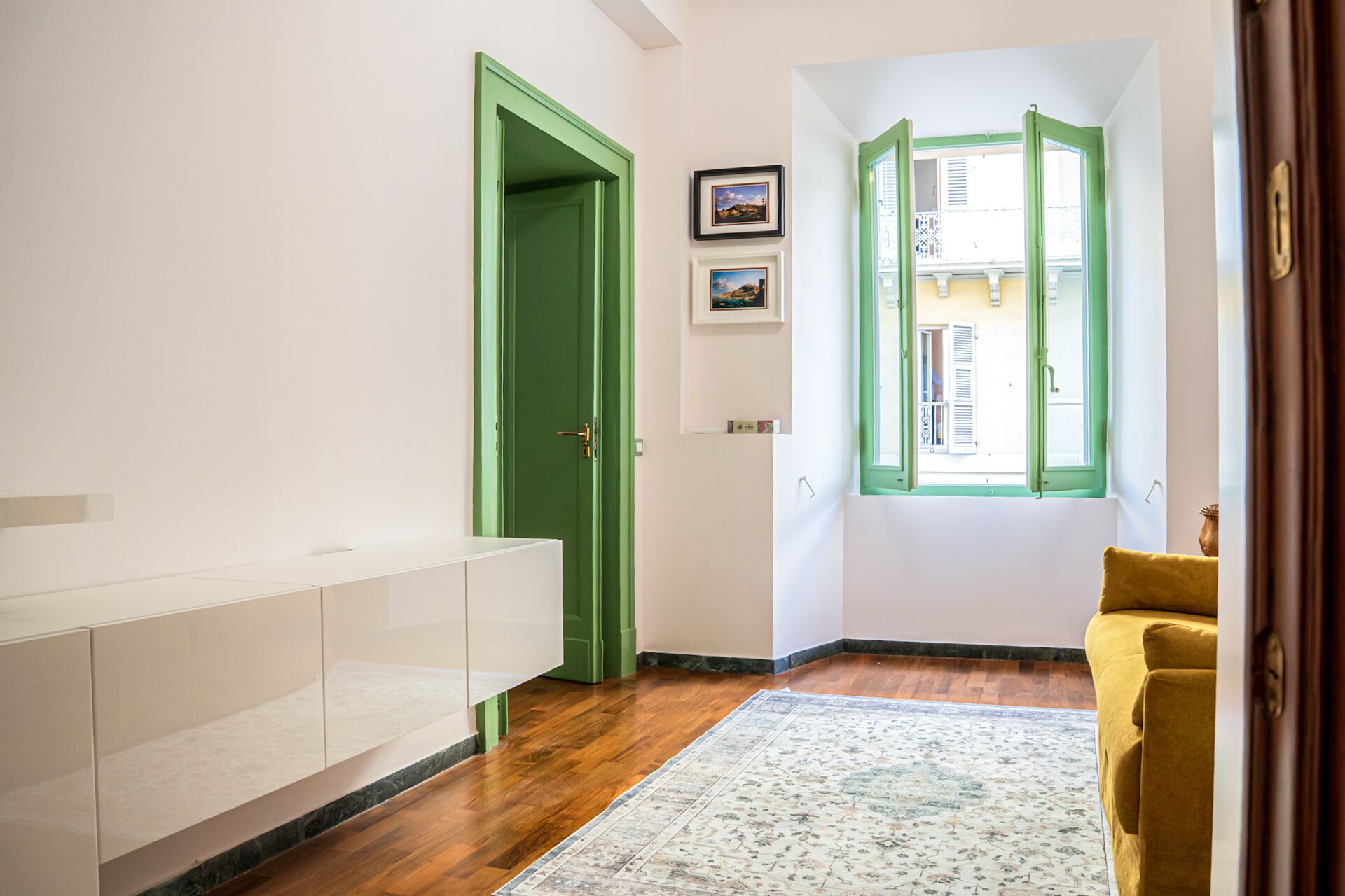 Un couloir avec une porte verte, une armoire blanche, un fauteuil jaune et une fenêtre donnant sur des immeubles.
