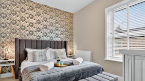 Ein Schlafzimmer mit gemustertem Wandpapier, einem gesteppten Kopfteil, einer grau gepolsterten Bank und einem Nachttisch mit Lampe und Tablett mit Frühstücksartikeln.