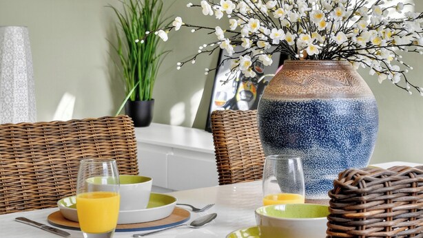 Ein gedeckter Esstisch mit Tellern, Gläsern und Besteck, ergänzt durch eine Vase mit weißen Blumen und eine Topfpflanze.