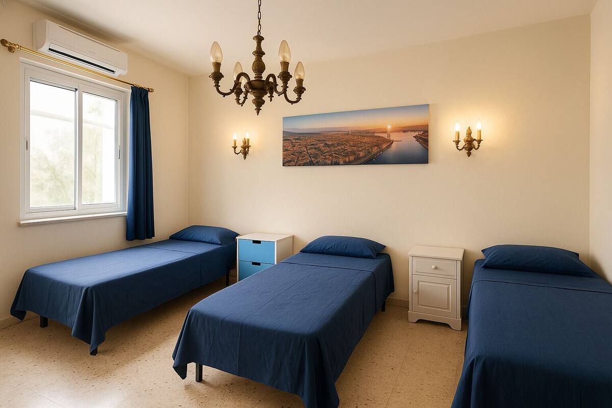 Ein Zimmer mit drei Einzelbetten, einem fensterbelaubten Fenster, einer Klimaanlage, einem Kronleuchter, Wandleuchten, einem Nachttisch, einer Schublade und einem Stadtbild.