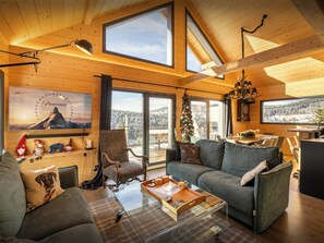 A cozy cabin interior with a wooden interior, a large window, a dining area, and a living room with a sofa and a glass coffee table.