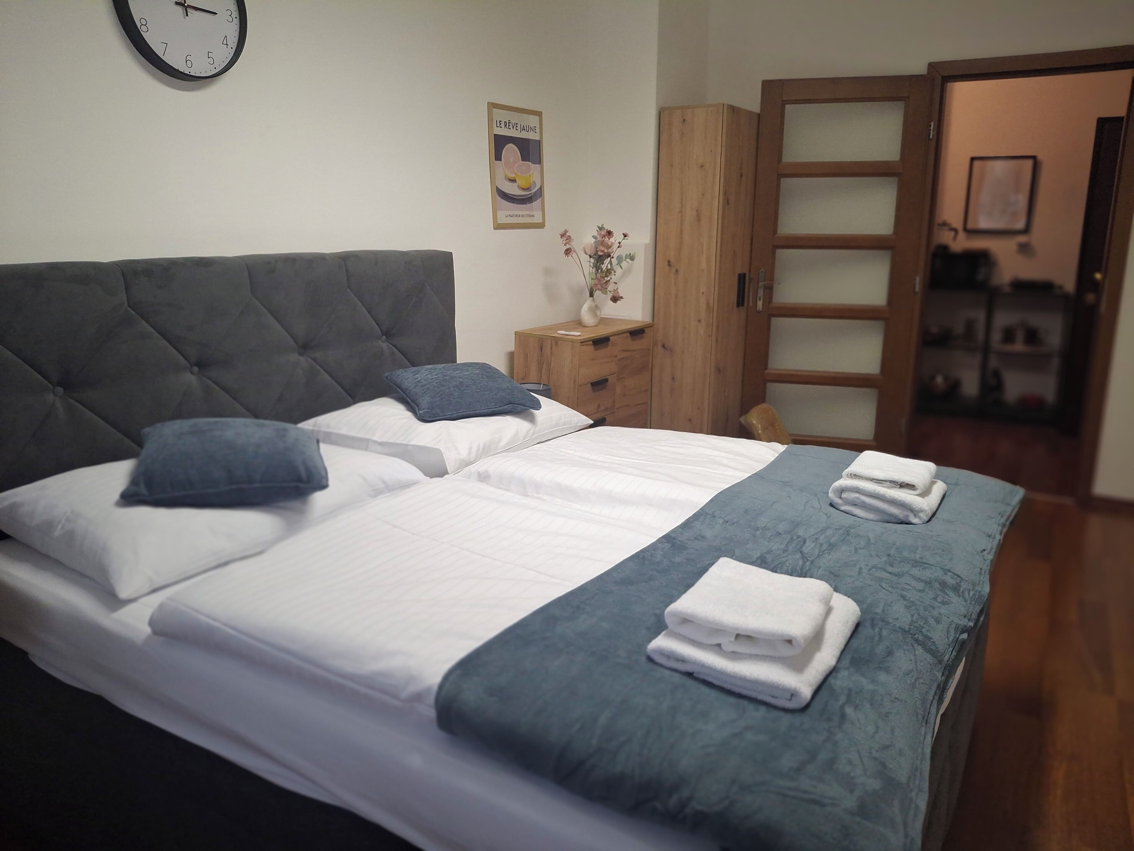 A neatly made bed with white linens, a grey headboard, and a blue accent pillow. There is a wooden nightstand with a vase of flowers beside the bed.