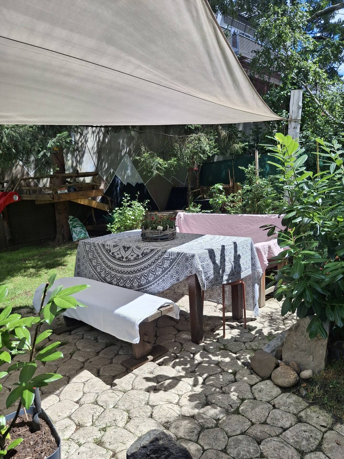 An outdoor patio with a table covered by a patterned tablecloth, a bench, and a canopy providing shade.