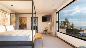 Una moderna camera d'albergo con un letto grande, un tavolino, una sedia, una TV e un'ampia finestra vista palme e mare.