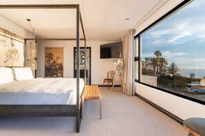 A modern hotel room with a large bed, a small table, a chair, a TV, and a large window with a view of palm trees and the sea. - Camps Bay Villa, Sleeps 10, Pool, Views - Cape House (Cape Town)
