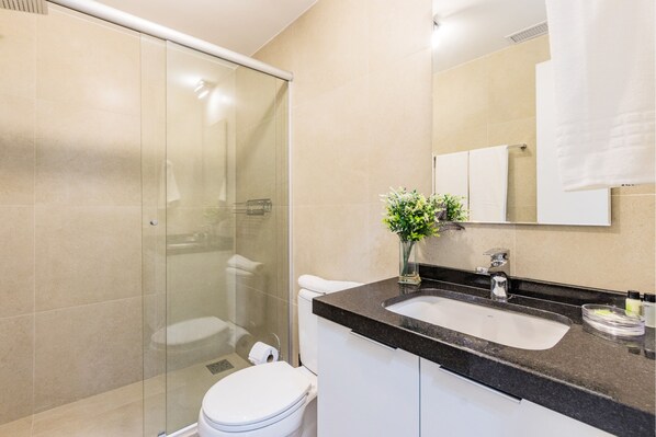 Ein Badezimmer mit einer gläsernen Duschkabine, einer weißen Toilette, einem Waschbecken mit Marmorplatte und einem Spiegel über dem Waschbecken.