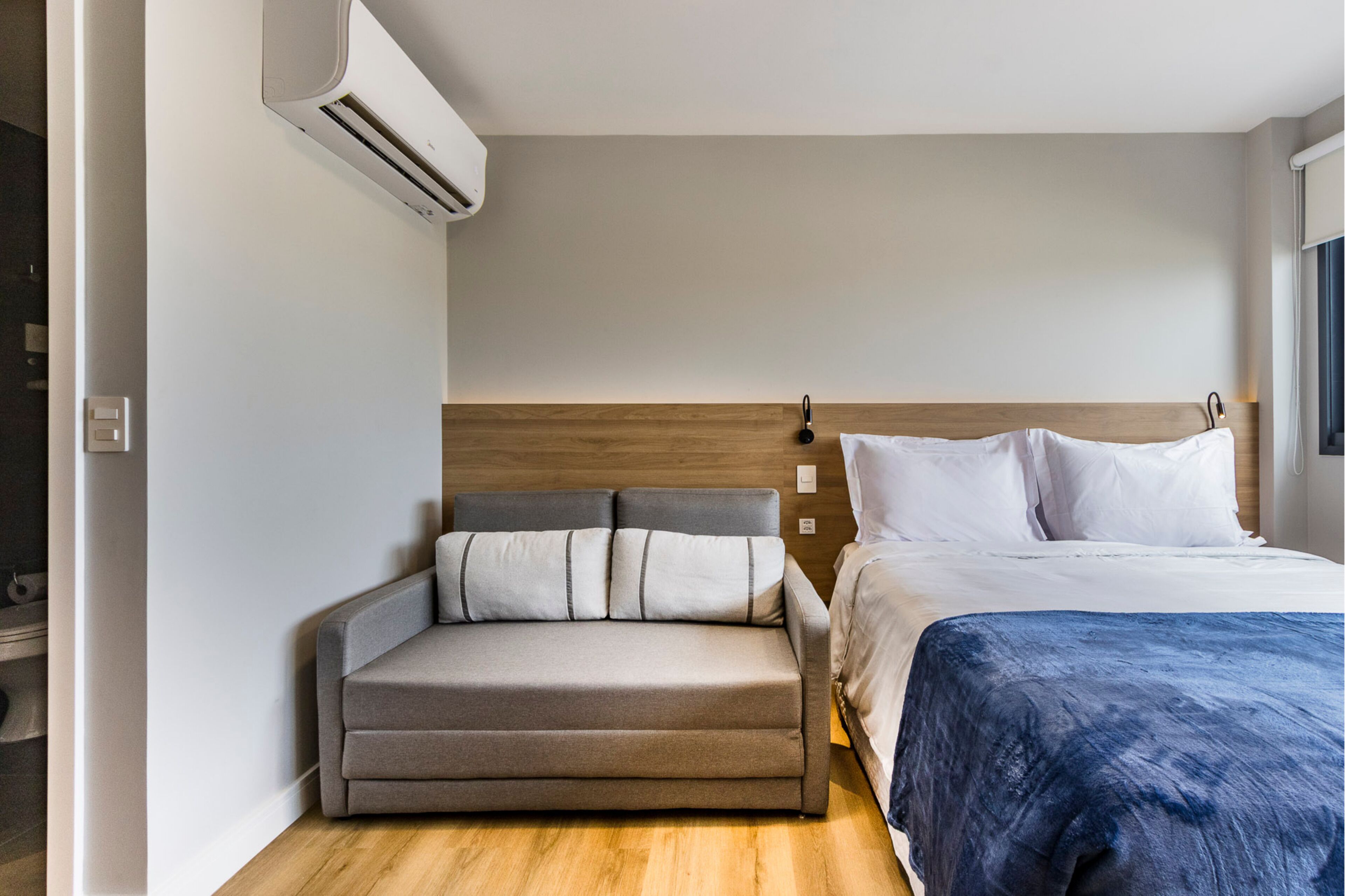 A hotel room with a bed, a sofa, a wooden headboard, and an air conditioning unit.