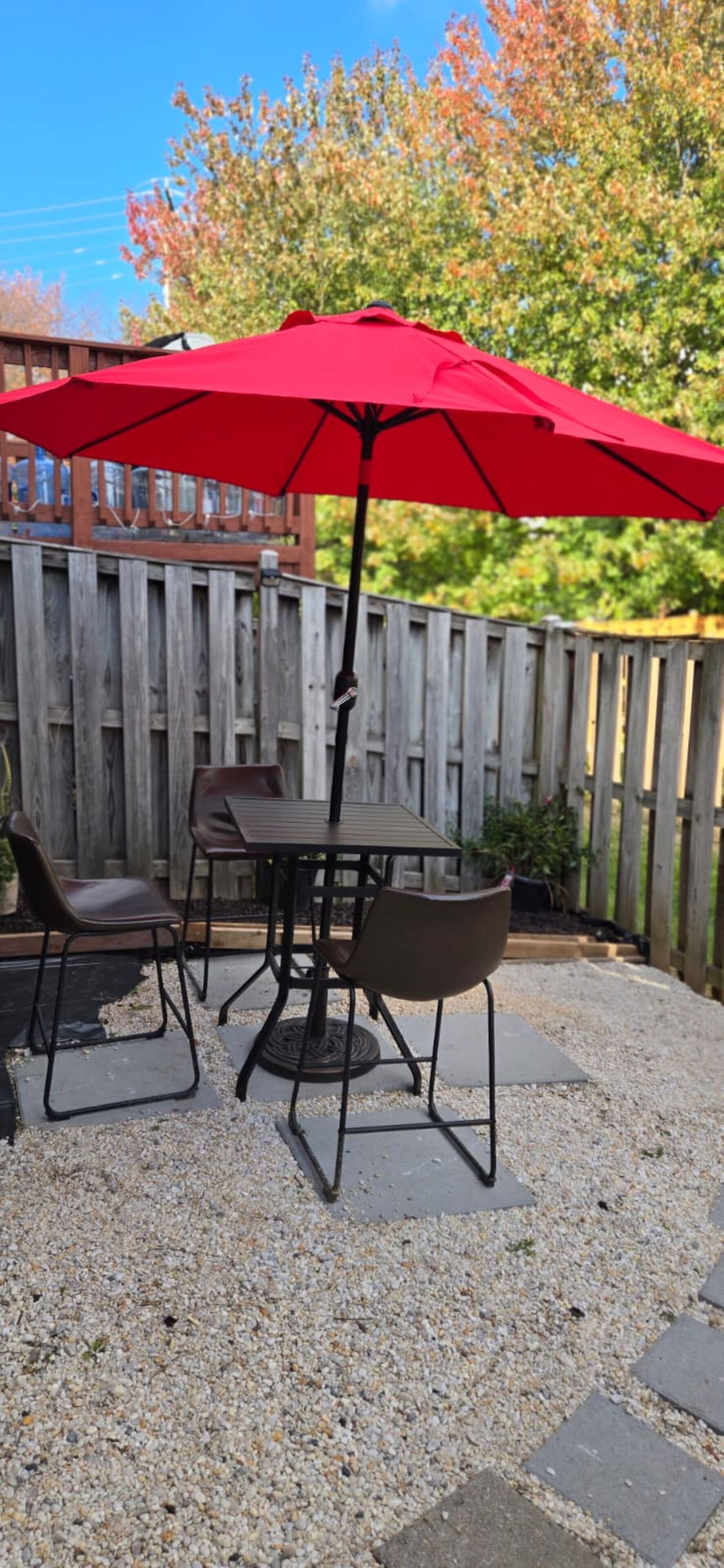 Une terrasse avec une table et des chaises sous un parasol rouge, entourée d’une clôture en bois et d’un sol en gravier.