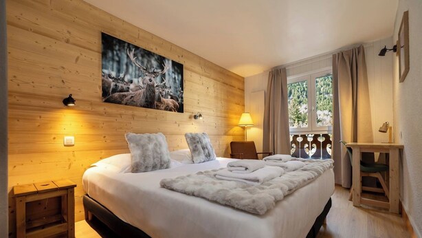 A cozy room with a large bed, wooden walls, a painting of deer, a window with curtains, and a small table. - Petit Social - Chaleureux | 4pax | Vue Montagne | Remontée300m (Chamonix-Mont-Blanc)