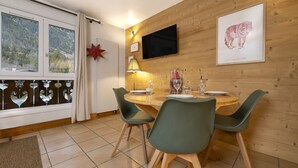 A dining area with a round wooden table, green chairs, a TV mounted on the wall, and a framed animal picture. - Petit Social - Chaleureux | 4pax | Vue Montagne | Remontée300m (Chamonix-Mont-Blanc)