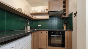 A compact kitchen with a dishwasher, oven, and wooden cabinets. - Petit Social - Chaleureux | 4pax | Vue Montagne | Remontée300m (Chamonix-Mont-Blanc)