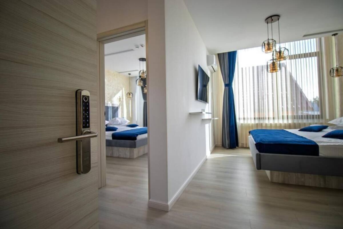 A modern hotel room with a wooden door, a bed with blue bedding, a second bed, a TV mounted on the wall, and a large window with curtains.