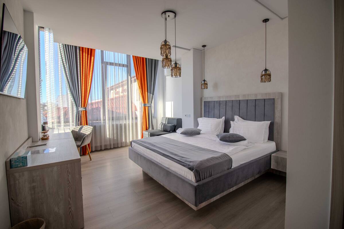 A modern bedroom with a large bed, a desk, and a view of buildings through sheer curtains.