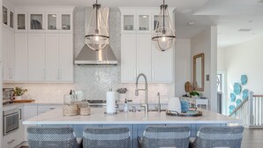 Une cuisine moderne avec des meubles blancs, un îlot central doté de tabourets de bar, un éclairage suspendu et un dosseret orné d’un motif en chevrons.