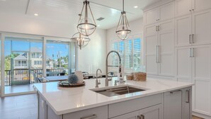 Une cuisine moderne avec des meubles blancs, un grand îlot central doté d’un évier, des luminaires suspendus et une vue sur une maison à travers de grandes fenêtres.