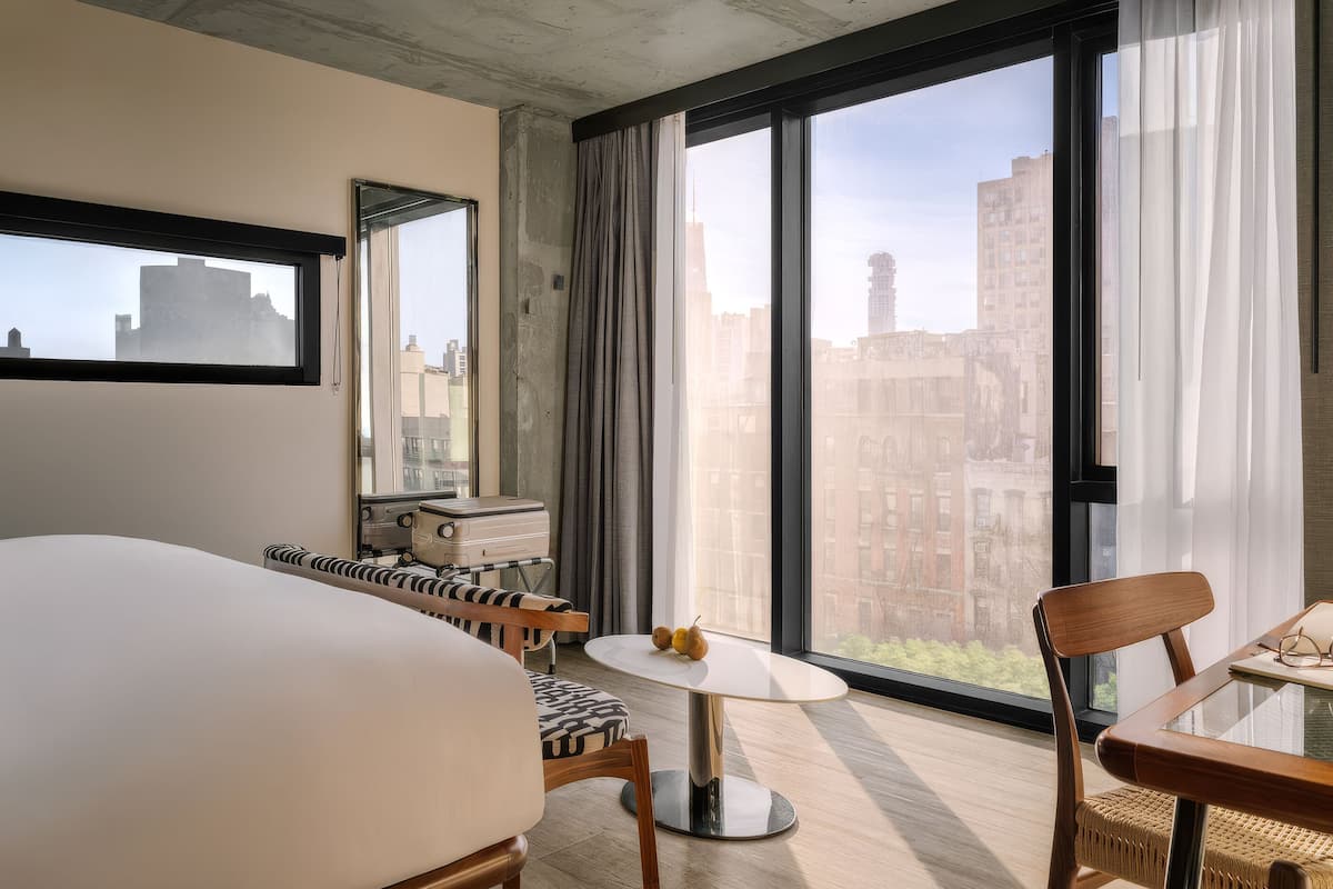 A hotel room with a large bed, a small table with a chair, a suitcase, and a view of the city through large windows.