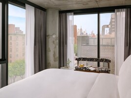A hotel room with a large bed, a view of the cityscape, and a small table with a book and a cup.