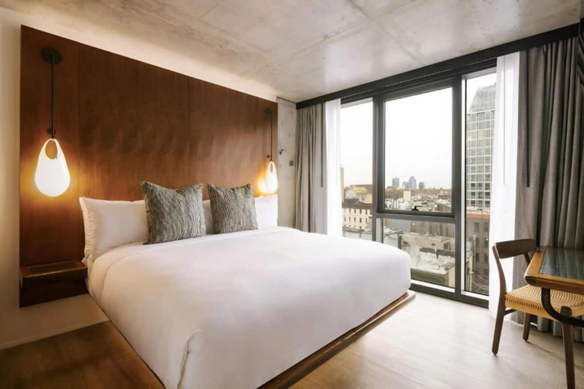 A hotel room with a large bed, a view of the city through large windows, and a wooden headboard.