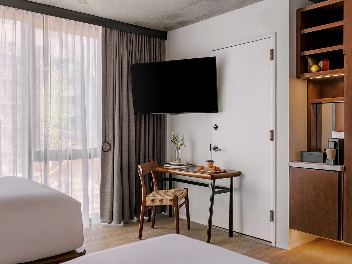 A hotel room with a bed, a desk with a chair, a television, a wardrobe, and a window with curtains.