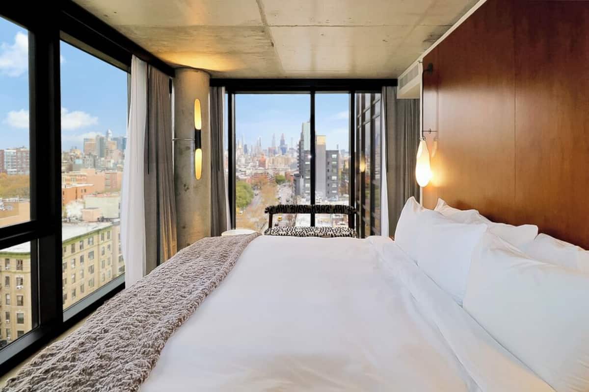 A hotel room with a large bed, a view of the city through floor-to-ceiling windows, and a city skyline in the background.