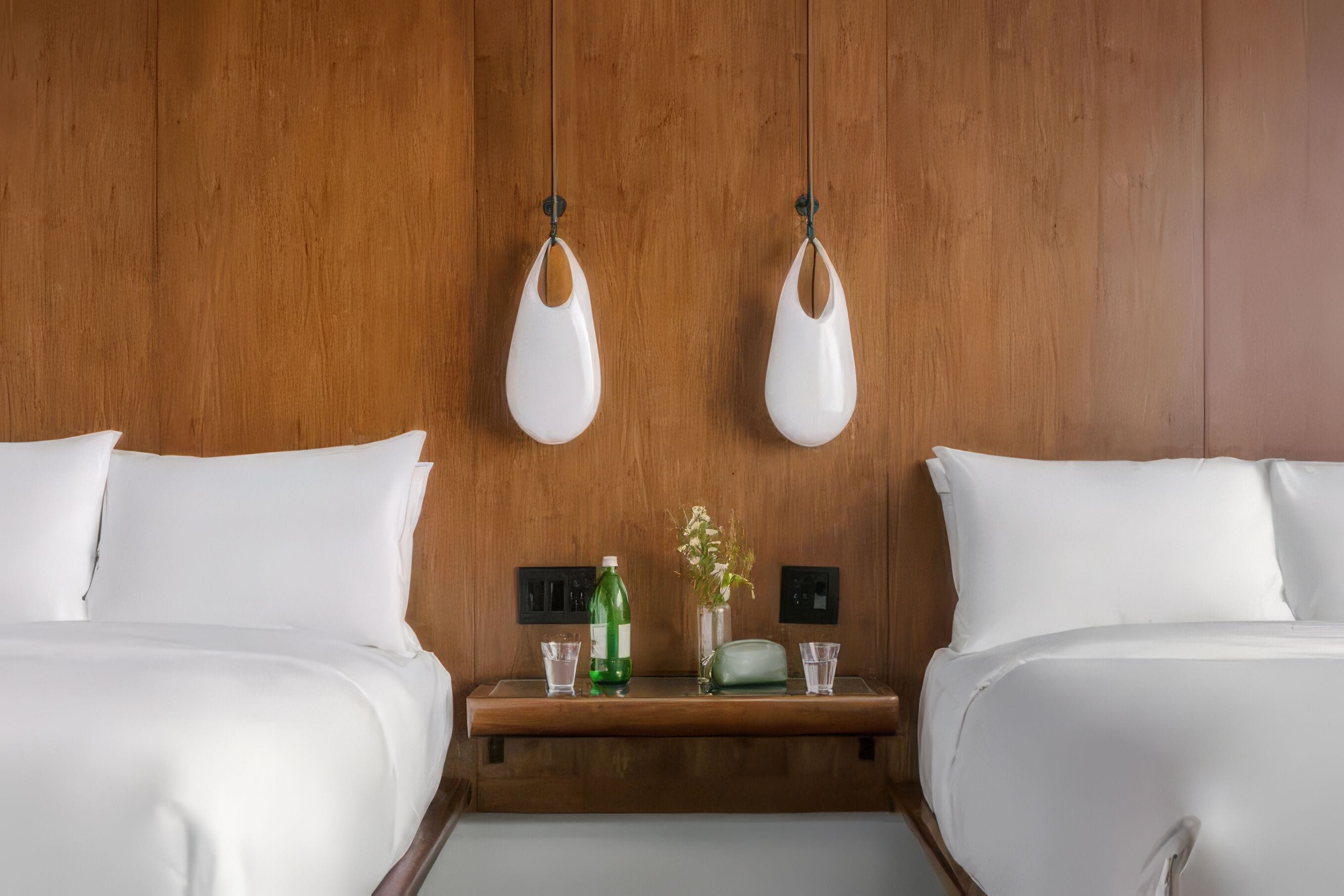 A hotel room with two beds, a wooden headboard, a small wooden nightstand with a green bottle, a glass, and a vase with flowers.