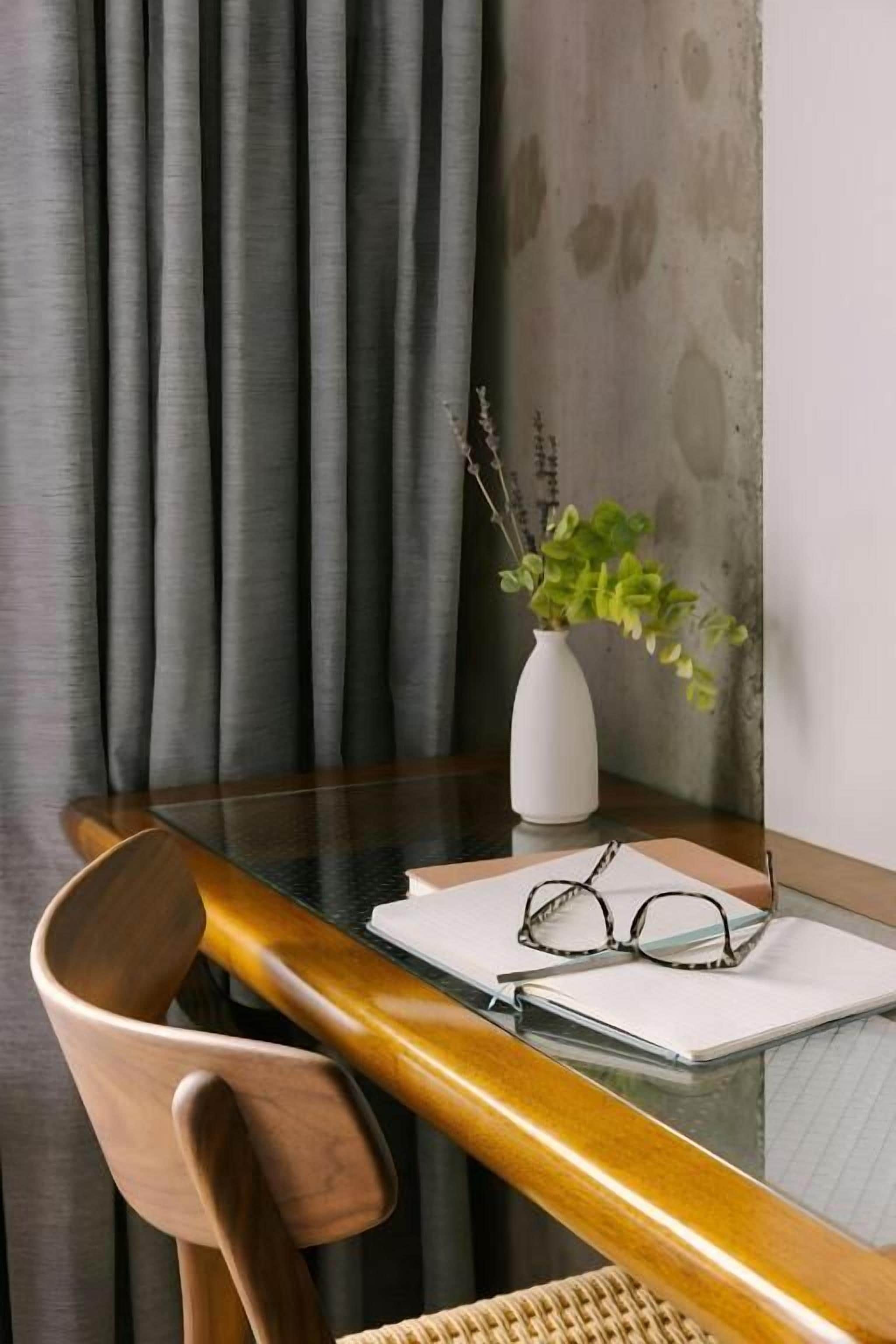 Een houten stoel, een kleine witte vaas met groene planten en een paar brillen op een glazen tafel.