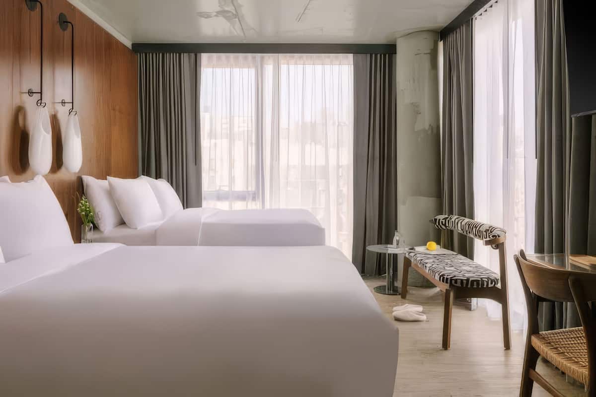 A hotel room with two beds, a small table, a chair, and a view of the city through large windows.