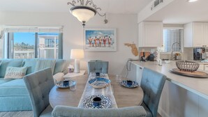 Un moderno salón con comedor, que incluye un sofá, una mesa preparada para dos personas y una cocina al fondo.