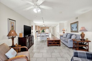A spacious living room with a sofa, a television, and a dining area in the background. - Superb 3-bedroom ocean view condo located within walking distance to everything! (Tybee Island)