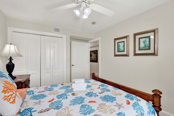 A bedroom with a bed, a nightstand, a lamp, a ceiling fan, and two framed pictures on the wall. - Superb 3-bedroom ocean view condo located within walking distance to everything! (Tybee Island)