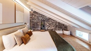 Une chambre avec un mur en pierre, un lit avec une literie blanche et marron, un plaid vert et une fenêtre donnant sur un beau paysage.