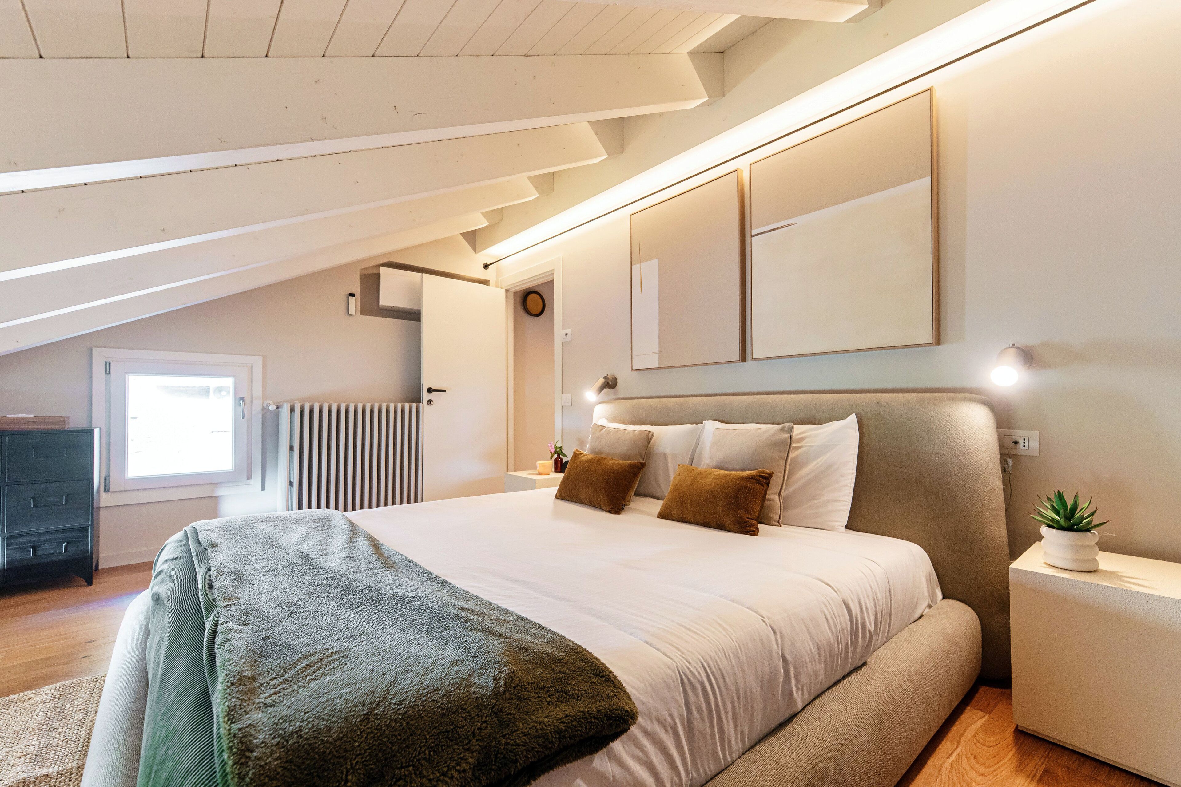 Een moderne slaapkamer met een groot bed, een nachtkastje met een plant en uitzicht op een raam en een radiator.