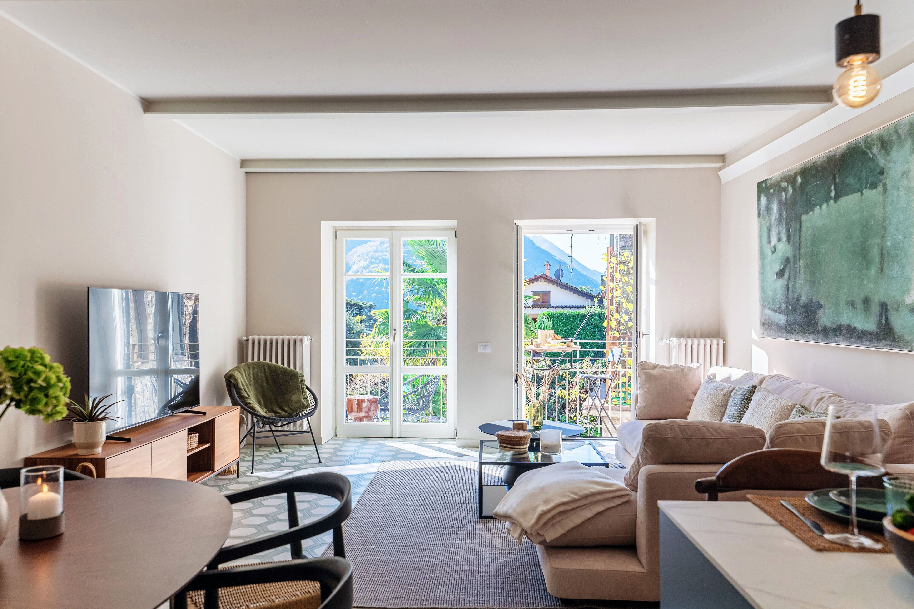 A spacious living room with a large TV, comfortable seating, a dining area, and a view through glass doors to a garden.