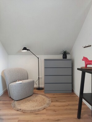 Una habitación de esquina con un sillón gris, una lámpara de pie, un armario gris y una pequeña mesa con un objeto rojo.