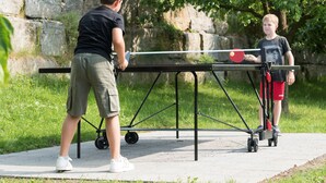 Zwei Personen spielen Tischtennis im Freien, ein Netz dazwischen, auf einem befestigten Untergrund, umgeben von Grünpflanzen und einer Steinmauer.