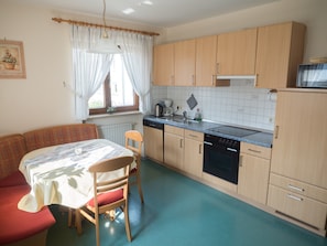 A compact kitchen with wooden cabinets, a dining table with chairs, and a window with curtains. - Holiday Farm Henn “Panorama” with Pool, Animals and Playground (Weikersheim)