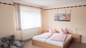 Ein Schlafzimmer mit einem Doppelbett, zwei Nachttischen, einem Schrank, einem Fenster mit Vorhängen, einem Heizkörper, einem Sofa und einer Wand mit einem Gemälde.