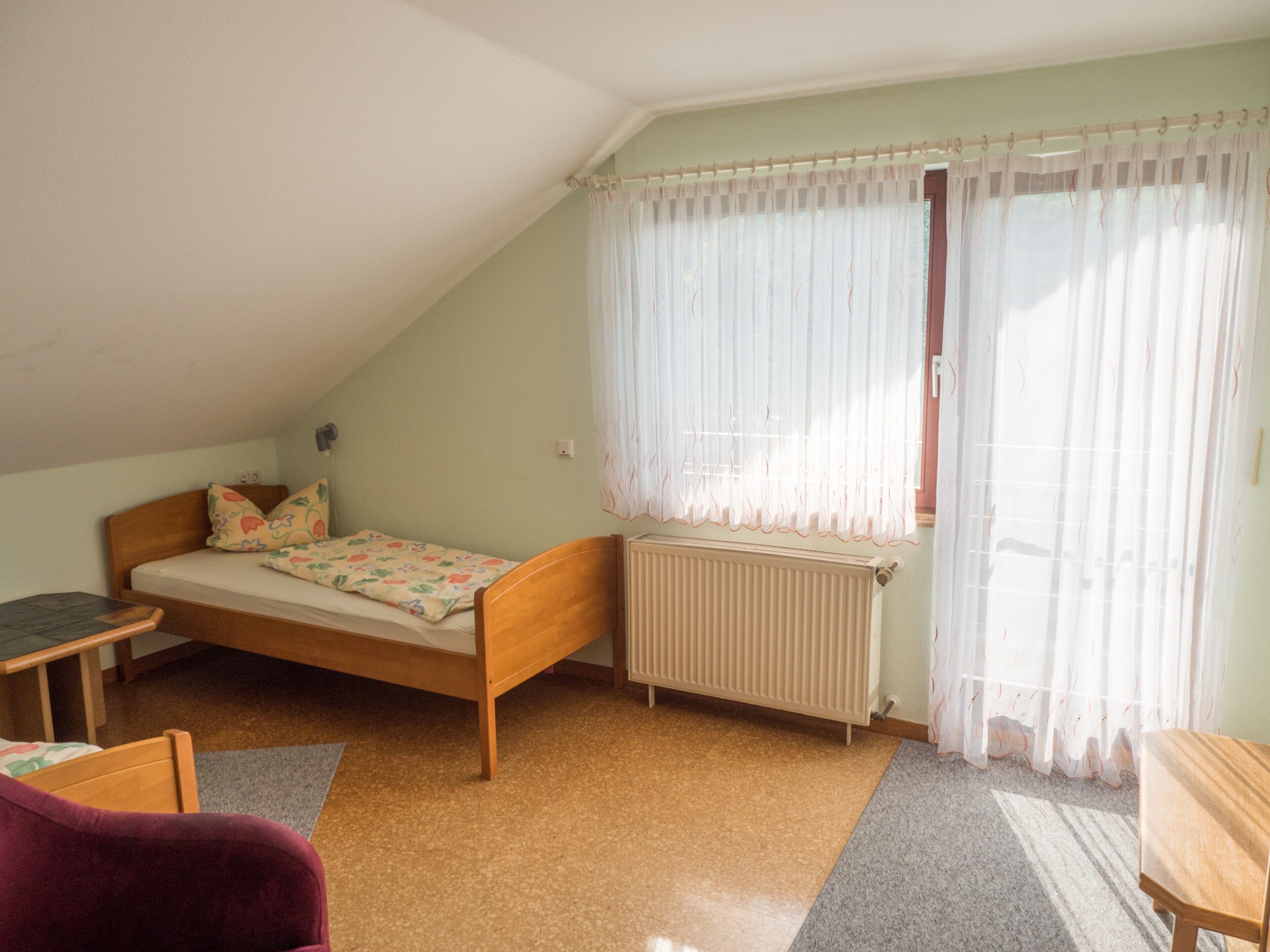 Un lit simple avec un cadre en bois, un radiateur, une fenêtre avec des rideaux légers et une petite table.