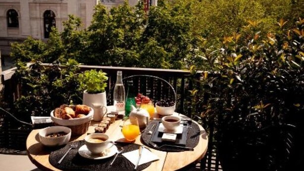 A round wooden table with breakfast items, a bottle, and a cup of coffee, set against a backdrop of lush greenery and a classical building. - Suite aux Anges N°3 (Duplex) (Antwerpen)