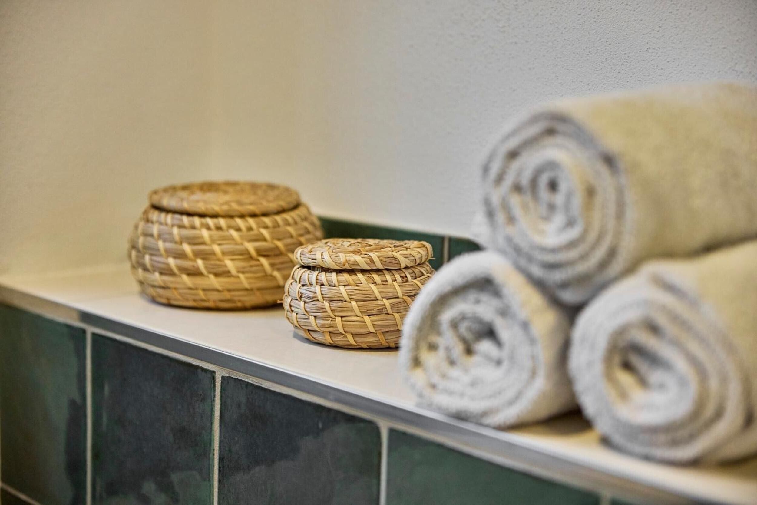 A bathroom with a tiled wall, a woven basket, and rolled towels.