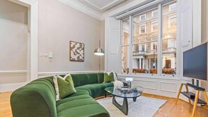 Ett vardagsrum med en grön soffa, ett soffbord i glas och ett stort fönster med utsikt över en byggnad.